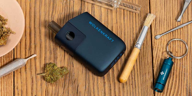 Schwarzer Vaporizer mit Cannabis-Blüten, Grinder und Zubehör auf Holztisch – Vorbereitung zum Vaporisieren statt Joint drehen