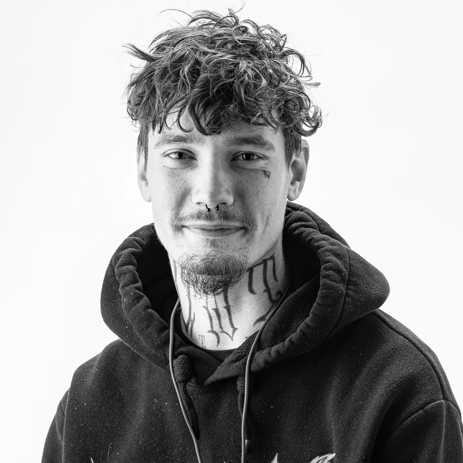 Teammitglied Marc – Smokee Aarau Porträt eines jungen Mannes mit lockigem Haar, Gesichtstattoos und Kapuzenpulli, vor hellem Hintergrund.